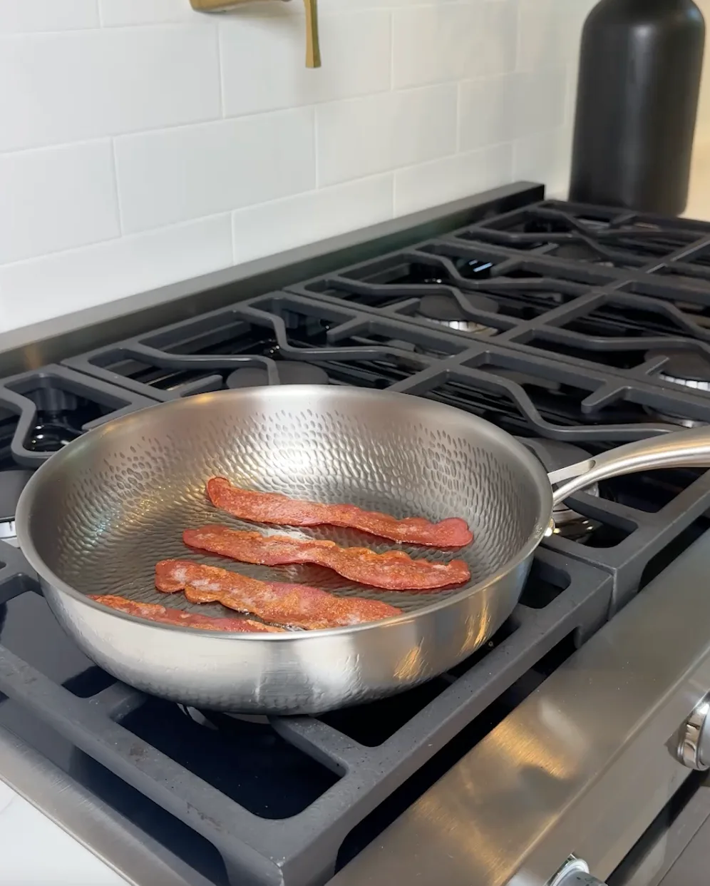 A titanium skillet with four strips of bacon cooking on a gas stovetop.